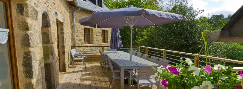 Terrasse en bois avec grande table, parasol et bacs fleuris le long de la façade en pierre du gîte