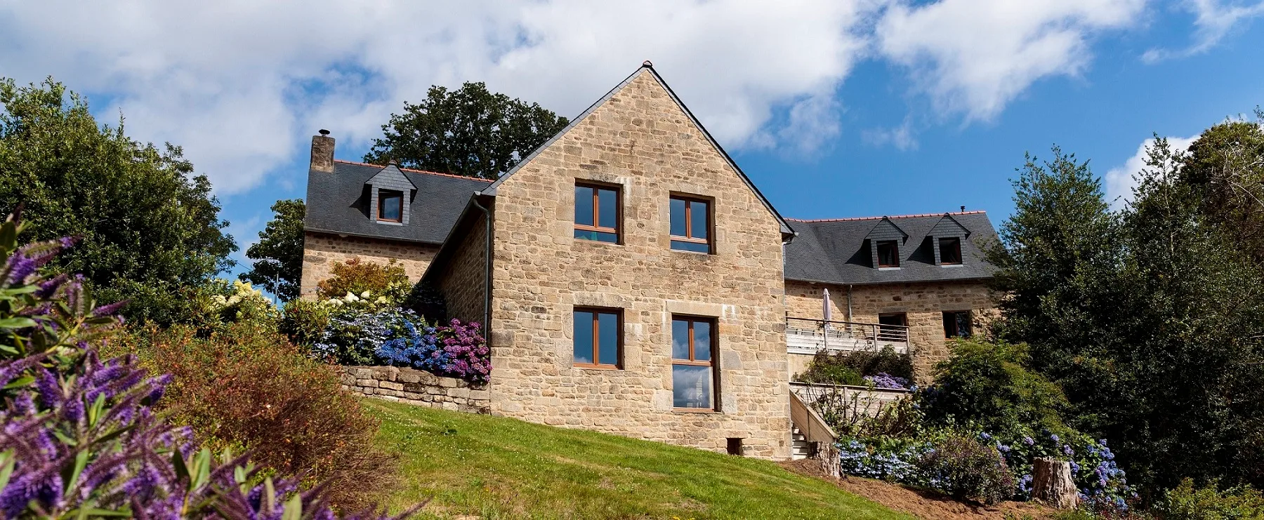 Gîte de groupe Le Vallon de Saint Eloi à Guiscriff — Grande maison en pierre à étage entourée d'un jardin fleuri d'hortensias sous un ciel bleu nuageux, capacité 15 personnes