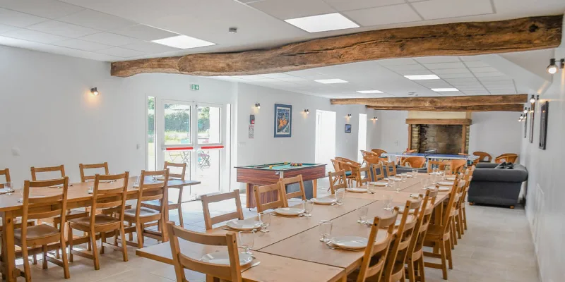 Grande salle de réception aux poutres apparentes, longues tables dressées, billard et cheminée en pierre