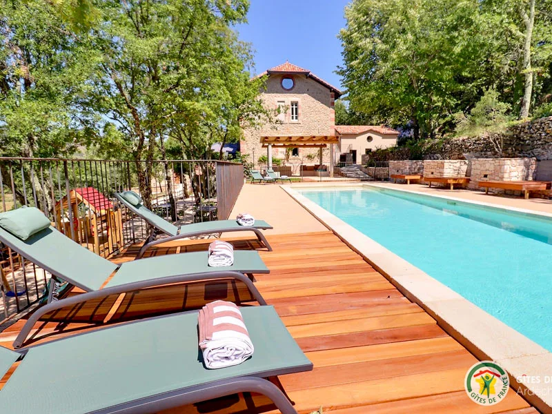 Piscine bordee de transats sur terrasse bois, maison en pierre et tonnelle en arriere-plan