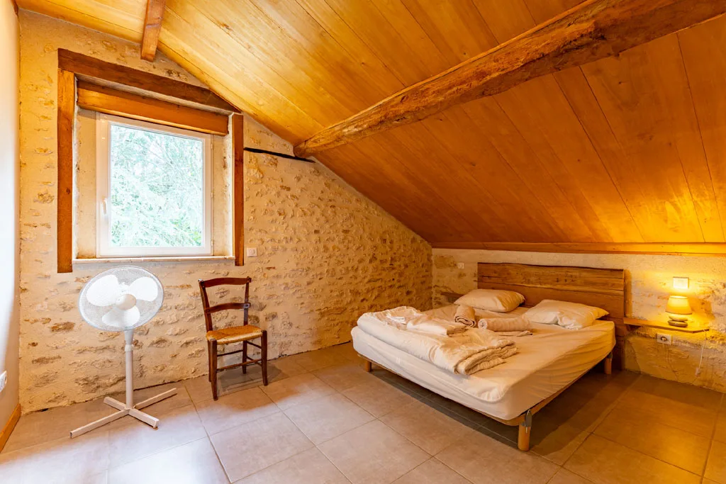 Chambre mansardée aux murs en pierre avec grand lit, chevet éclairé et fenêtre sur la verdure