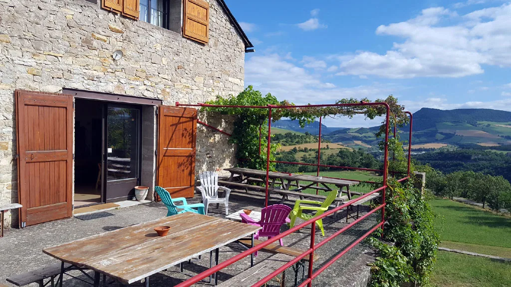 Gîte de groupe Gîte de Cabrières à Compeyre — Grande terrasse extérieure avec longues tables, bancs colorés et tonnelle fleurie ouverte sur les collines, capacité 18 personnes