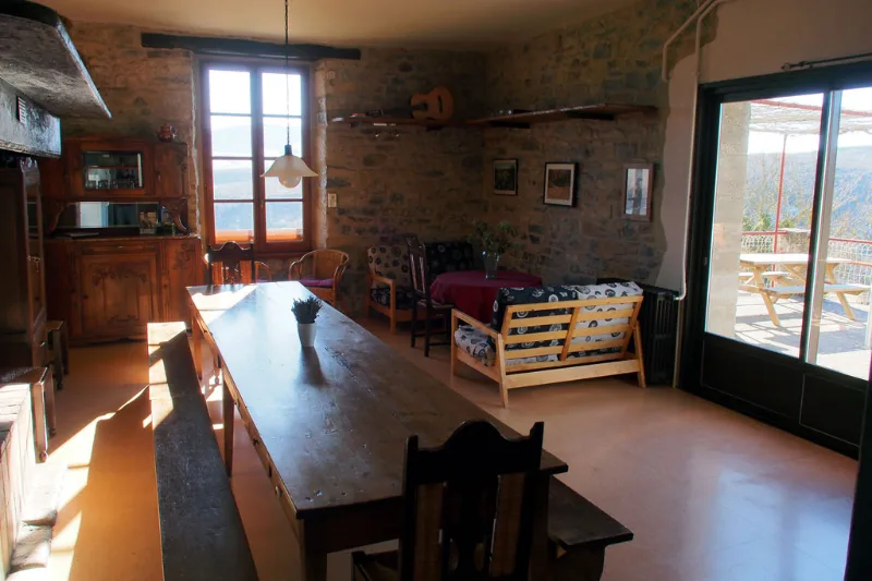 Grand séjour en pierre avec longue table en bois, banquettes et baie vitrée vers la terrasse