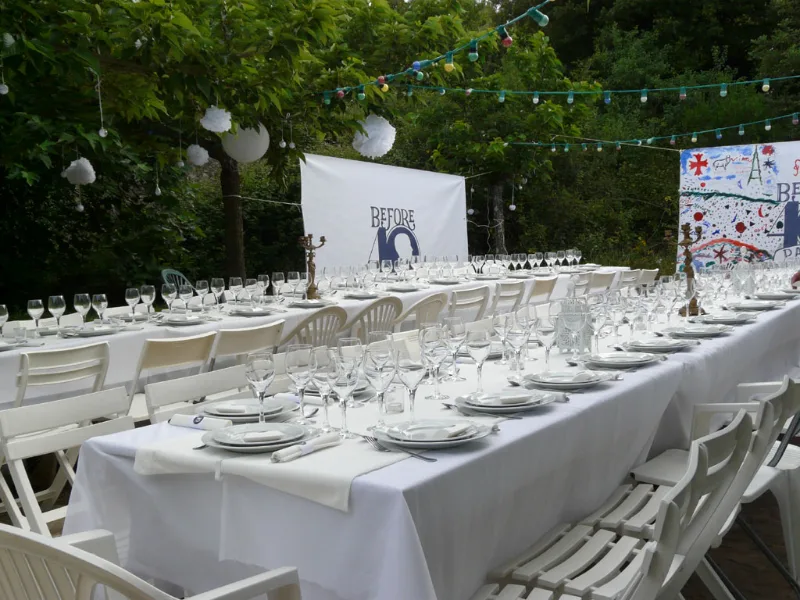 Longues tables nappées de blanc dressées sous les arbres avec guirlandes lumineuses pour fête
