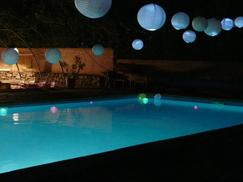 Piscine éclairée la nuit avec lampions suspendus et boules lumineuses flottant sur l'eau