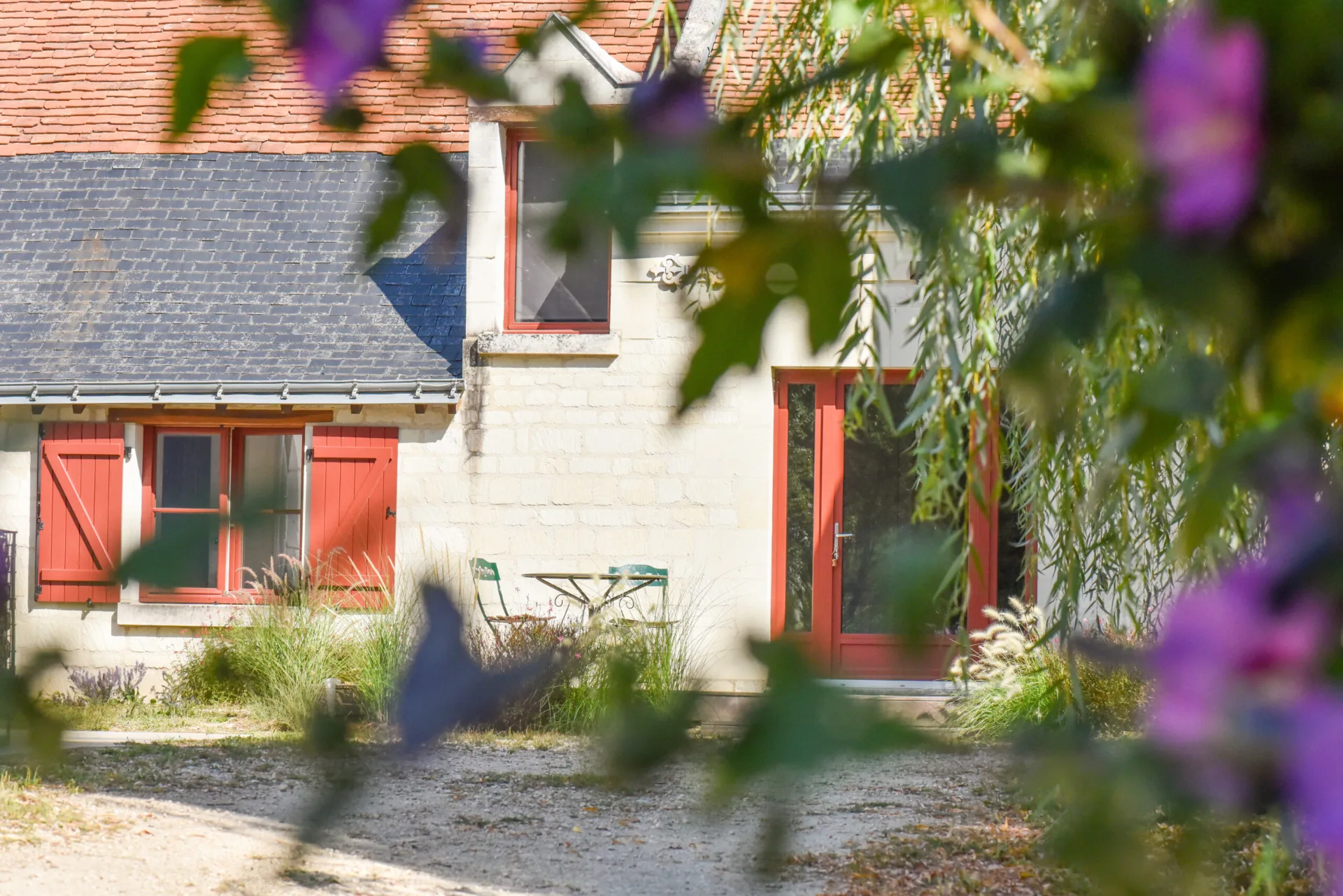 Gîte de groupe La Maison de Thélème à Huismes — Longère en tuffeau aux volets rouges, avec porte vitrée ouverte sur la cour du domaine viticole, capacité 12 personnes