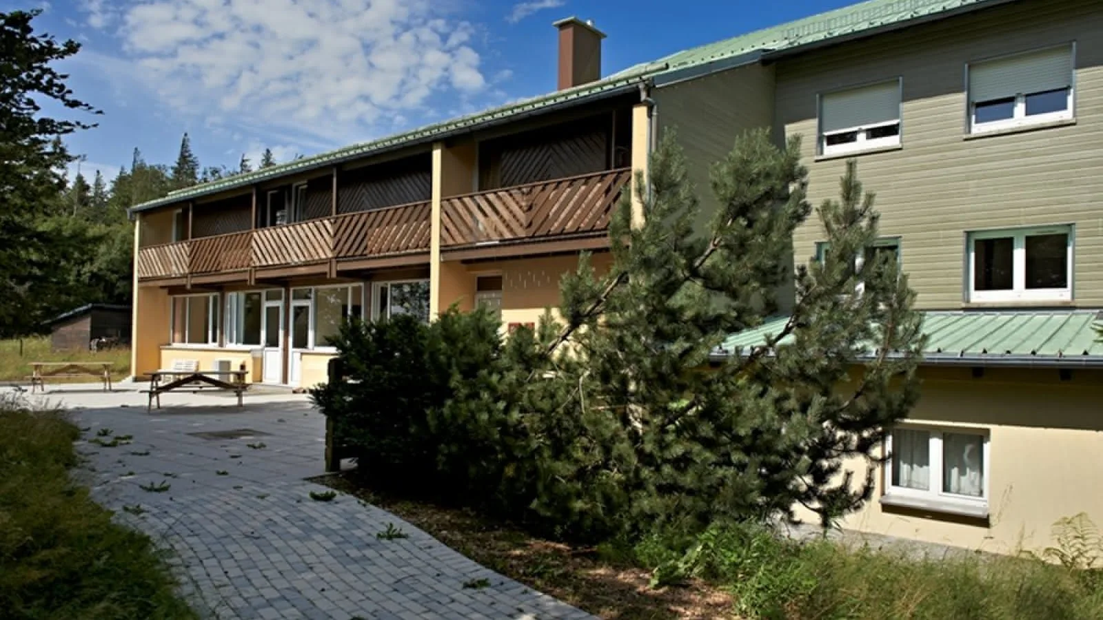 Gîte de groupe Les Sapins à Belmont — Façade extérieure du centre Les Sapins à Belmont, terrasse pavée avec tables de pique-nique entourée de sapins, capacité 45 personnes