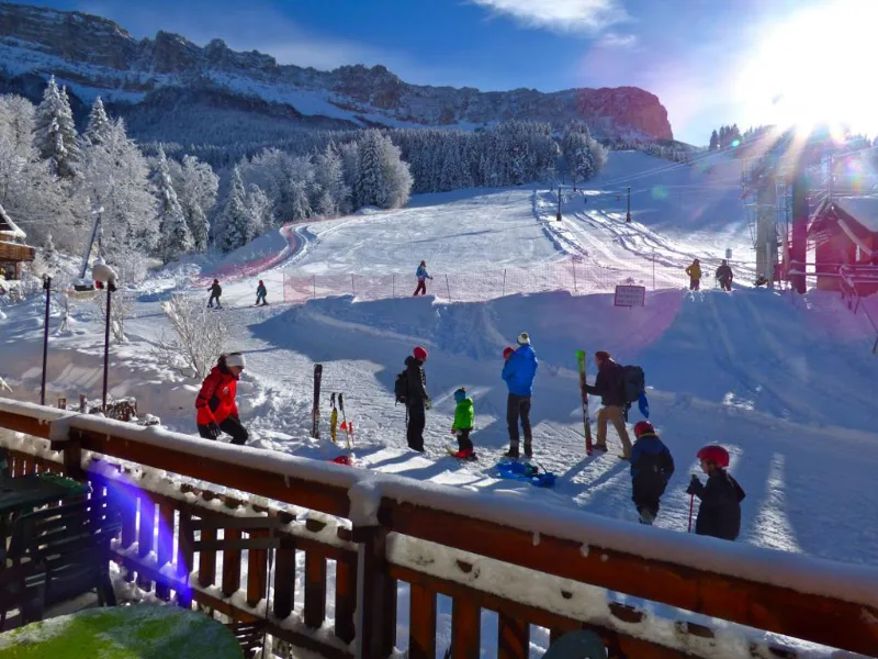 Vue depuis balcon sur piste de ski avec skieurs et massif rocheux enneigé au soleil