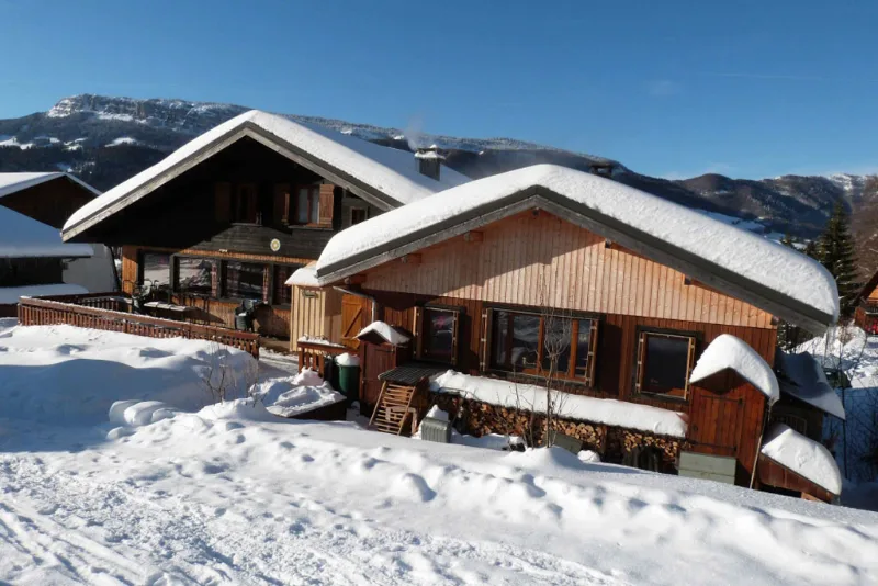 Deux chalets en bois enneigés avec réserve de bois et massif alpin sous ciel bleu