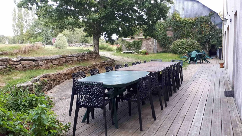 Grande terrasse en bois avec longues tables dressées pour un groupe, jardin arboré et dépendance en pierre