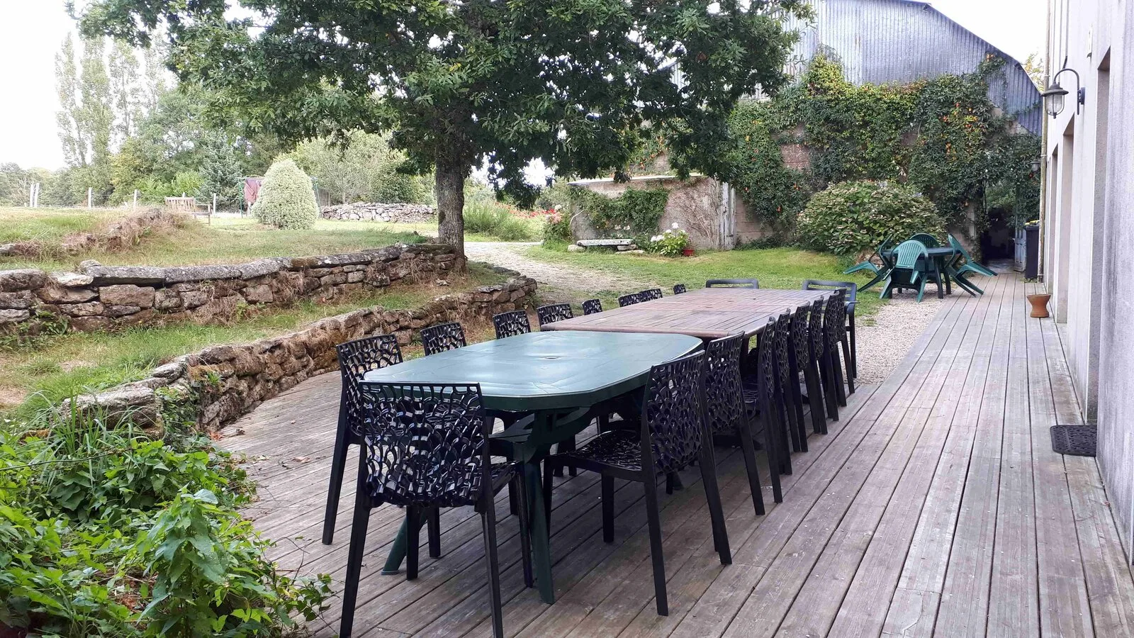 Gîte de groupe Grand Gîte Saint Colomban à Saint Nolff — Grande terrasse en bois avec longues tables dressées pour un groupe, jardin arboré et dépendance en pierre, capacité 15 personnes