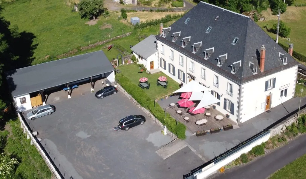 Gîte de groupe Le Clos Auvergnat à Tauves — Vue aérienne du corps de ferme auvergnat avec cour, terrasses ombragées et grand parking privé, capacité 49 personnes