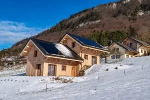 Deux chalets en bois avec panneaux solaires sous la neige, entourés de montagnes en hiver