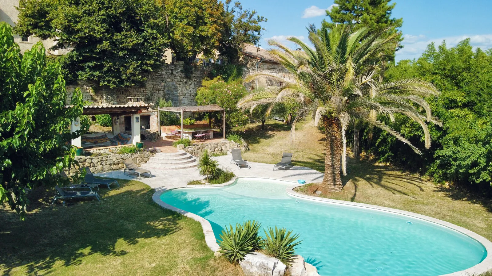 Gîte de groupe Mas du Côté Sud à Martignargues — Vue aérienne du mas en pierre avec piscine sinueuse entourée de palmiers et jardin méditerranéen, capacité 14 personnes