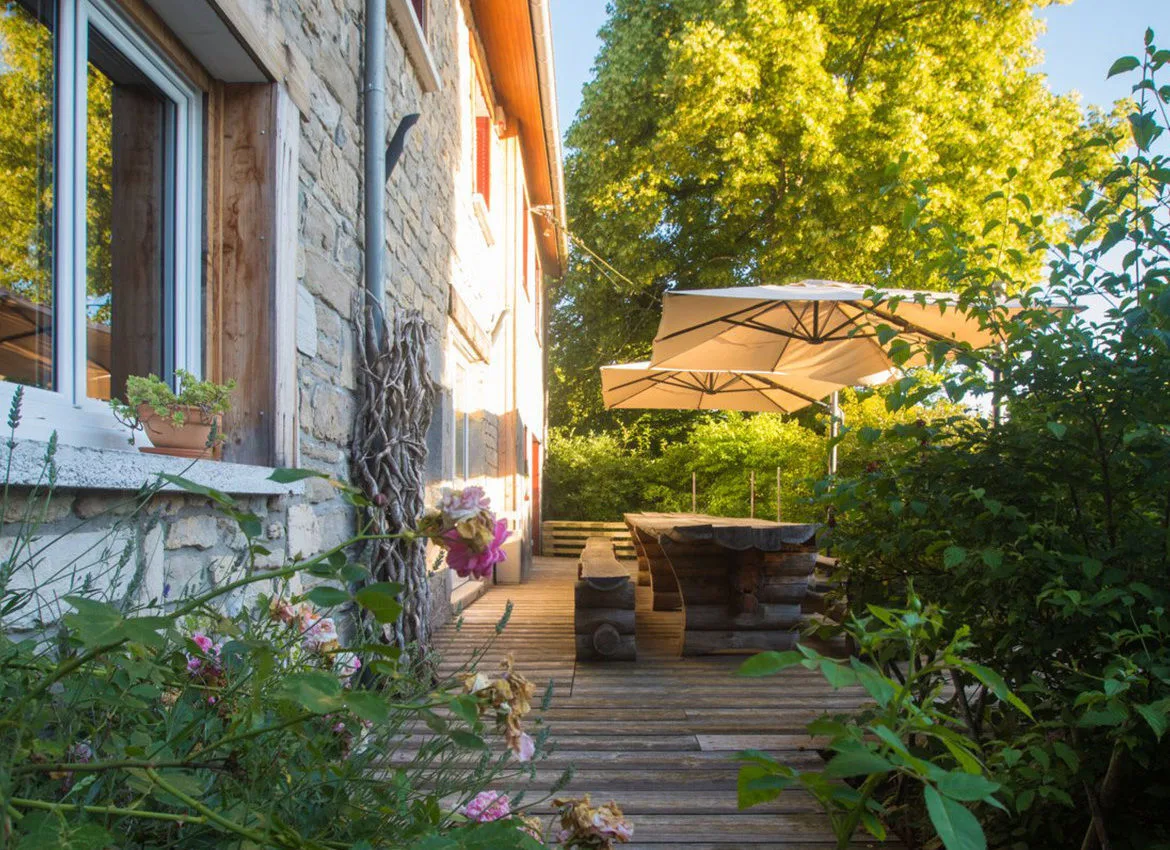 Gîte de groupe Les Terrasses de Merlue à Plaisia — Terrasse en bois ombragée devant la ferme en pierre avec grande table extérieure et parasols, capacité 26 personnes