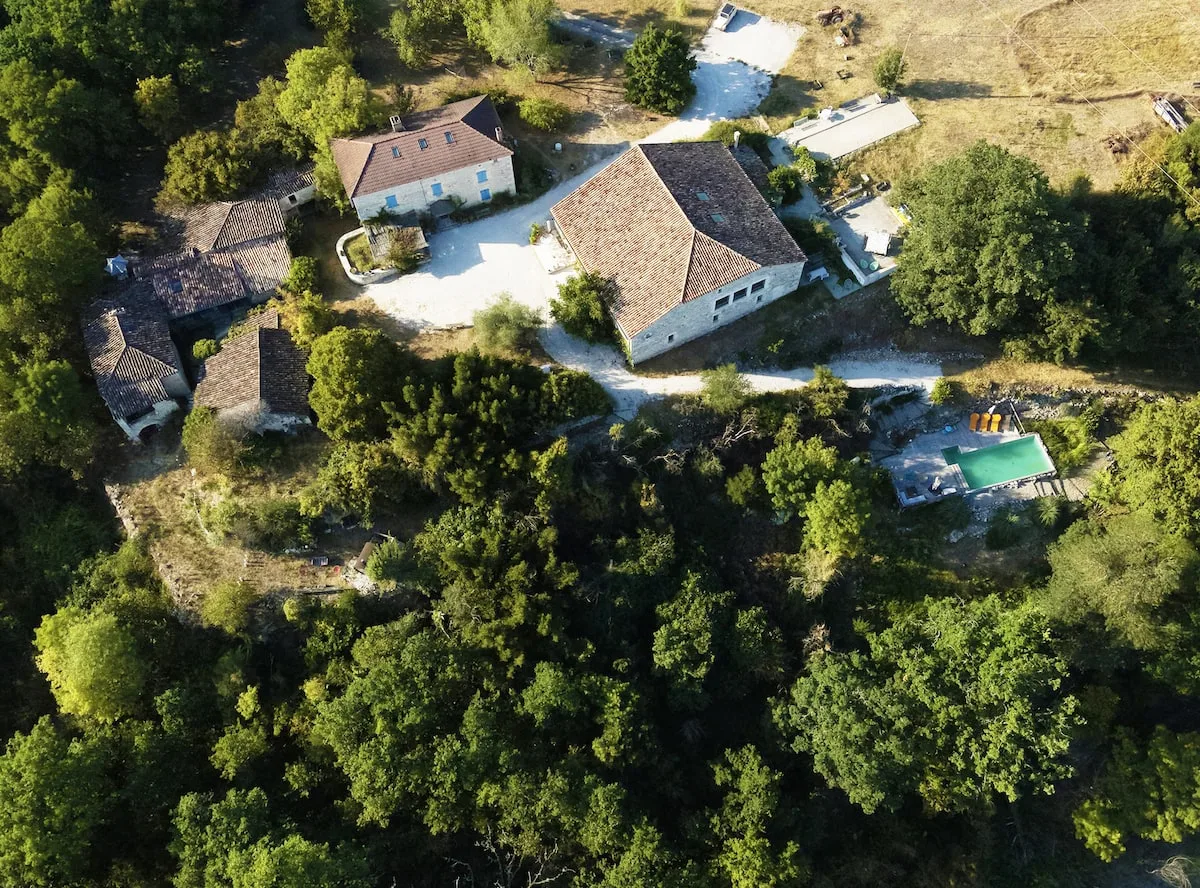 Gîte de groupe The Happy Hamlet à Fauroux — Vue aérienne du hameau en pierre avec toitures en tuiles et piscine entourée d'arbres, capacité 15 personnes