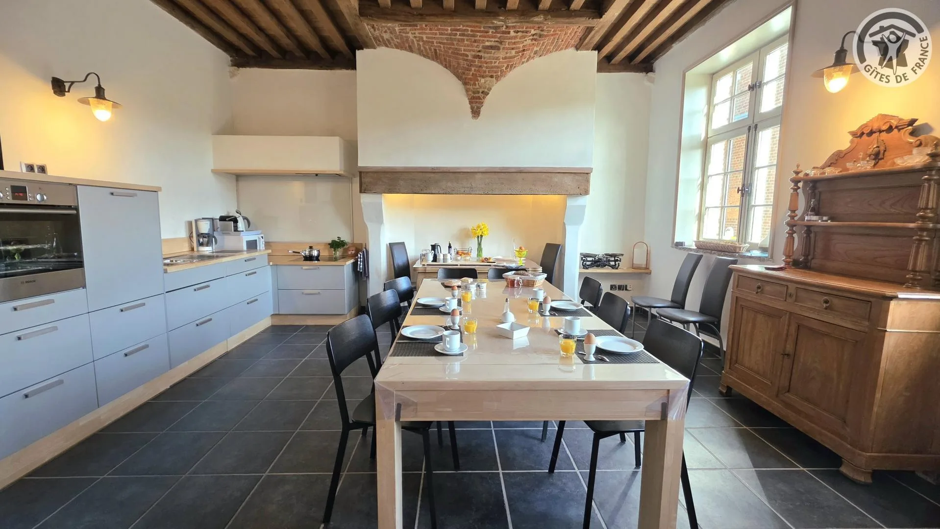 Cuisine ouverte avec grande table dressée pour le petit-déjeuner sous poutres et cheminée voûtée en brique