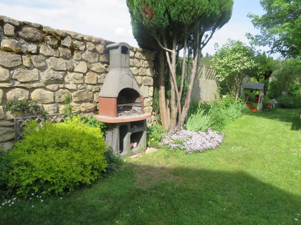 Barbecue en pierre dans un coin de jardin ombragé, entouré d'arbustes fleuris