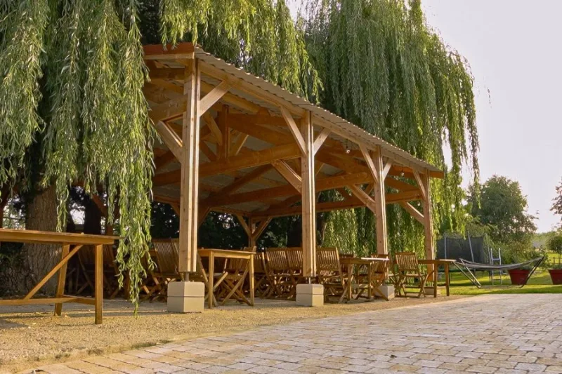 Grande pergola en bois couverte avec tables et bancs, installée sous les saules pour les repas de groupe
