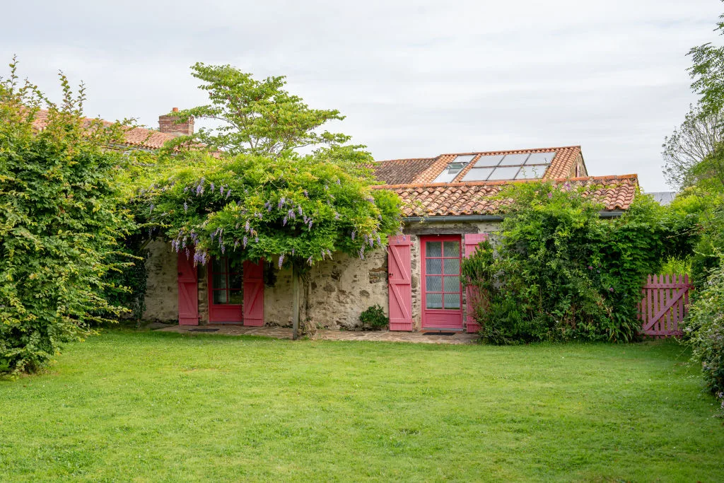 Façade arrière du gîte couverte de glycine en fleurs, portes rouges donnant sur le jardin