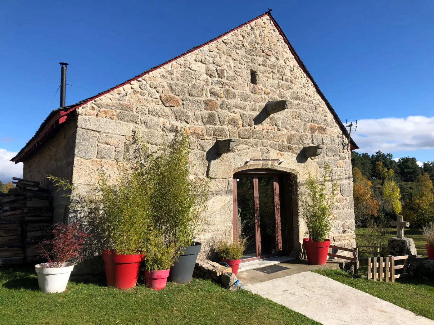 Gîte de groupe La Grange de Grandviala à Peyre en Aubrac — Façade en pierre de l'ancienne grange avec toit à double pente, terrasse fleurie et pots de bambous, capacité 15 personnes