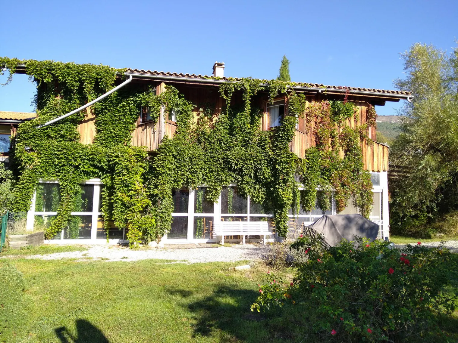 Gîte de groupe La Ferme du Chevalet à Aspres-sur-Buëch — Façade en bois et pierre couverte de vigne vierge, grande véranda vitrée au rez-de-chaussée, capacité 15 personnes