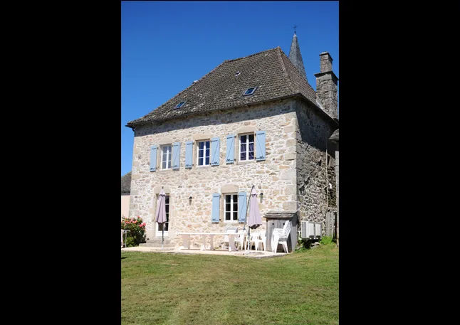 Façade en pierre avec volets bleus, terrasse aménagée avec table, chaises et jardin ombragé