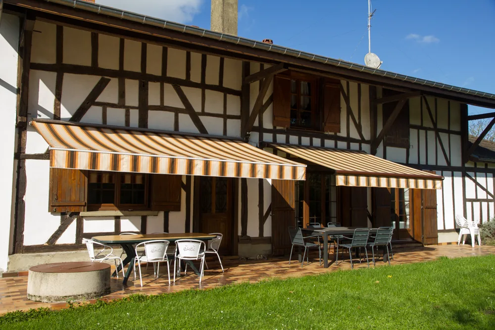 Gîte de groupe Gîte Rural des Trois Sillons à Louze — Façade à pans de bois du gîte avec terrasse couverte, tonnelle et mobilier de jardin, capacité 12 personnes