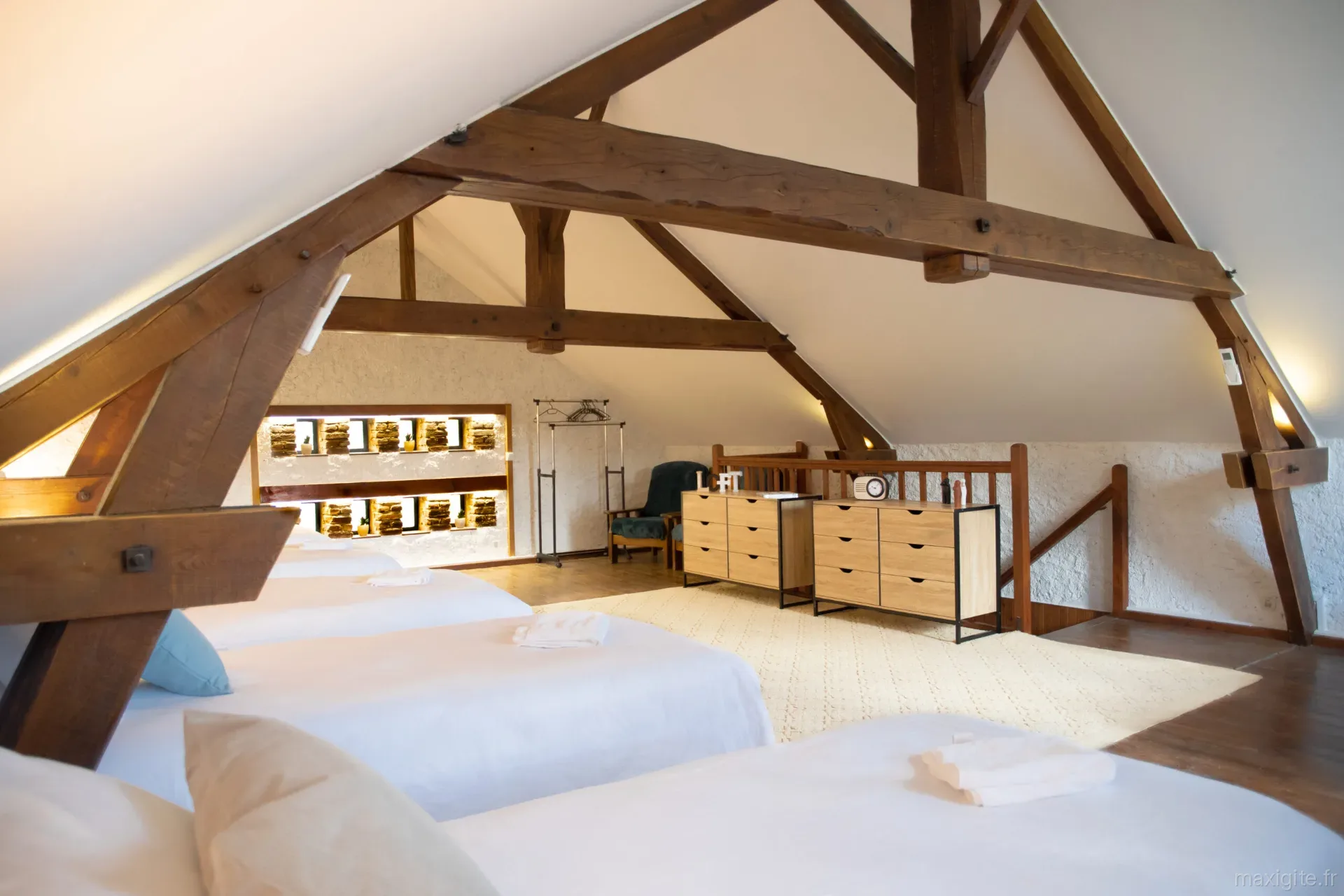 Grand dortoir sous charpente en bois avec plusieurs lits simples alignés et tapis clair