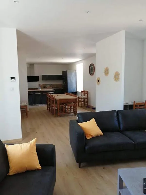 Grand séjour lumineux avec canapés noirs, coussins jaunes et coin repas en bois