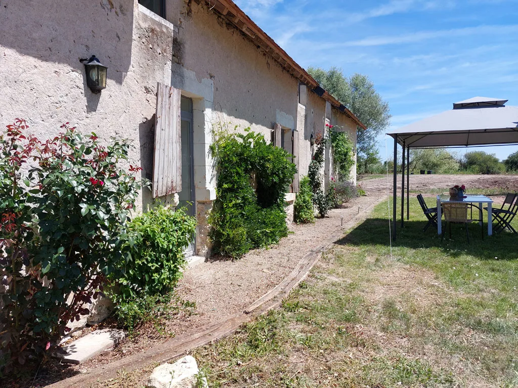 Façade en pierre du gîte avec roseraie, volets bois et coin repas sous tonnelle