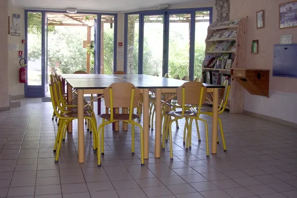 Grande salle commune avec longue table et chaises jaunes, baies vitrées sur la nature