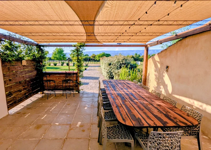 Terrasse couverte sous pergola avec grande table en bois et vue sur les champs et le Luberon