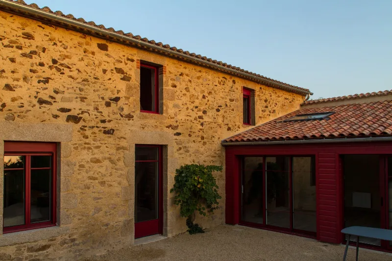 Façade en pierre du gîte avec volets rouges, terrasse et jardin fleuri