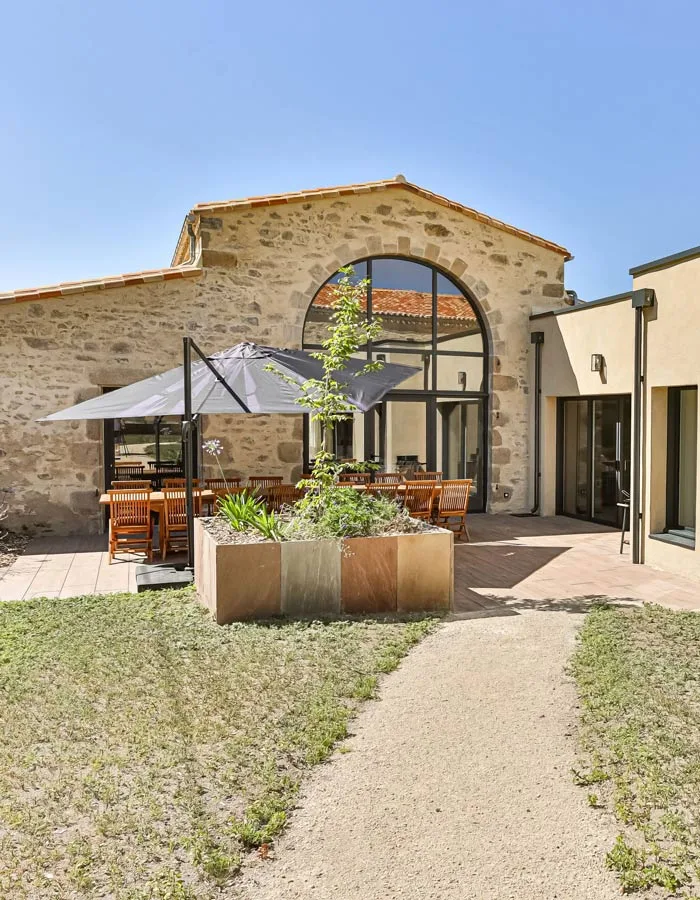 Gîte de groupe Gîtes des Pâturages à La Gaubretière — Façade en pierre du domaine avec terrasse ombragée, table en bois et grande baie vitrée, capacité 29 personnes