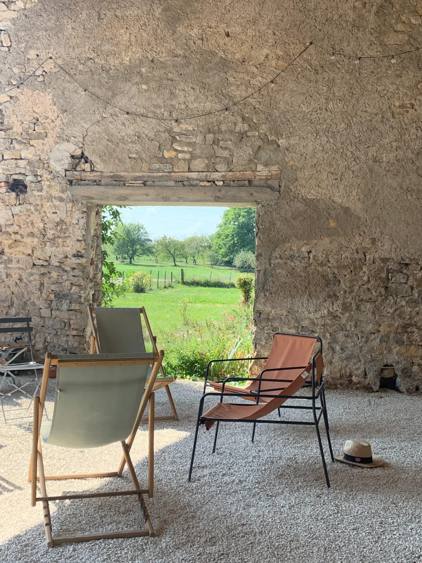 Gîte de groupe Le Clos des Maix à Bezange-la-Grande — Façade en pierre du Clos des Maix vue à travers une ouverture voûtée, jardin verdoyant et chaises longues, capacité 15 personnes