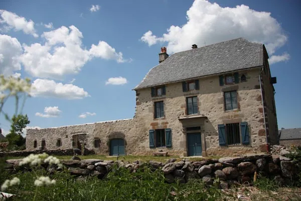 Gîte de groupe Fortuniès 1864 à Dienne — Façade en pierre de Fortuniès avec toit d'ardoises et volets bleus, ciel dégagé, capacité 20 personnes