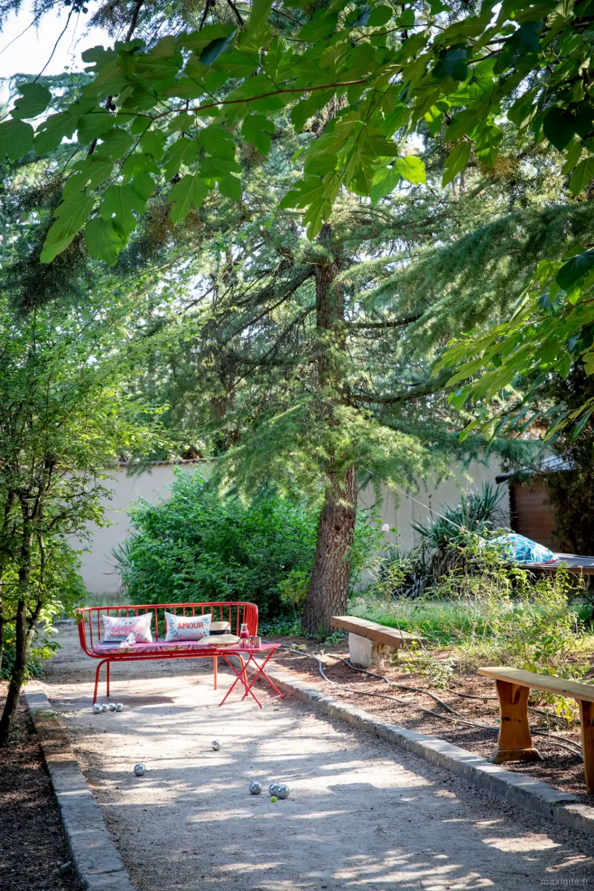 Allée de boulodrome ombragée par les pins, avec banc rouge et boules de pétanque