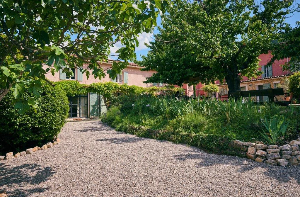Allée de gravier menant au bâtiment d'accueil avec jardin fleuri et arbres ombragés