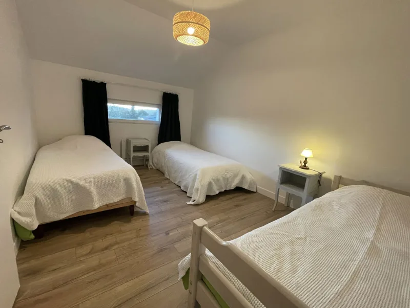 Chambre avec trois lits simples, parquet clair, rideaux noirs et luminaire tressé