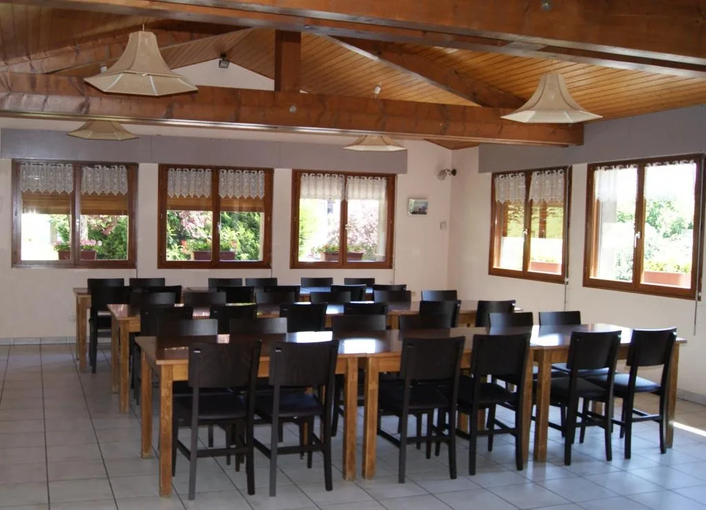 Salle de restauration avec plafond bois a poutres apparentes, tables en bois et chaises noires, suspensions lumineuses et fenetres sur jardin