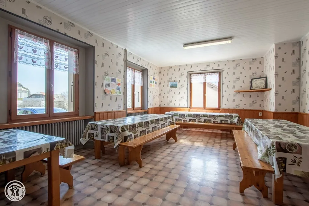 Salle d activites avec tables et bancs en bois, nappes a motifs, papier peint fleuri et sol en carrelage losanges