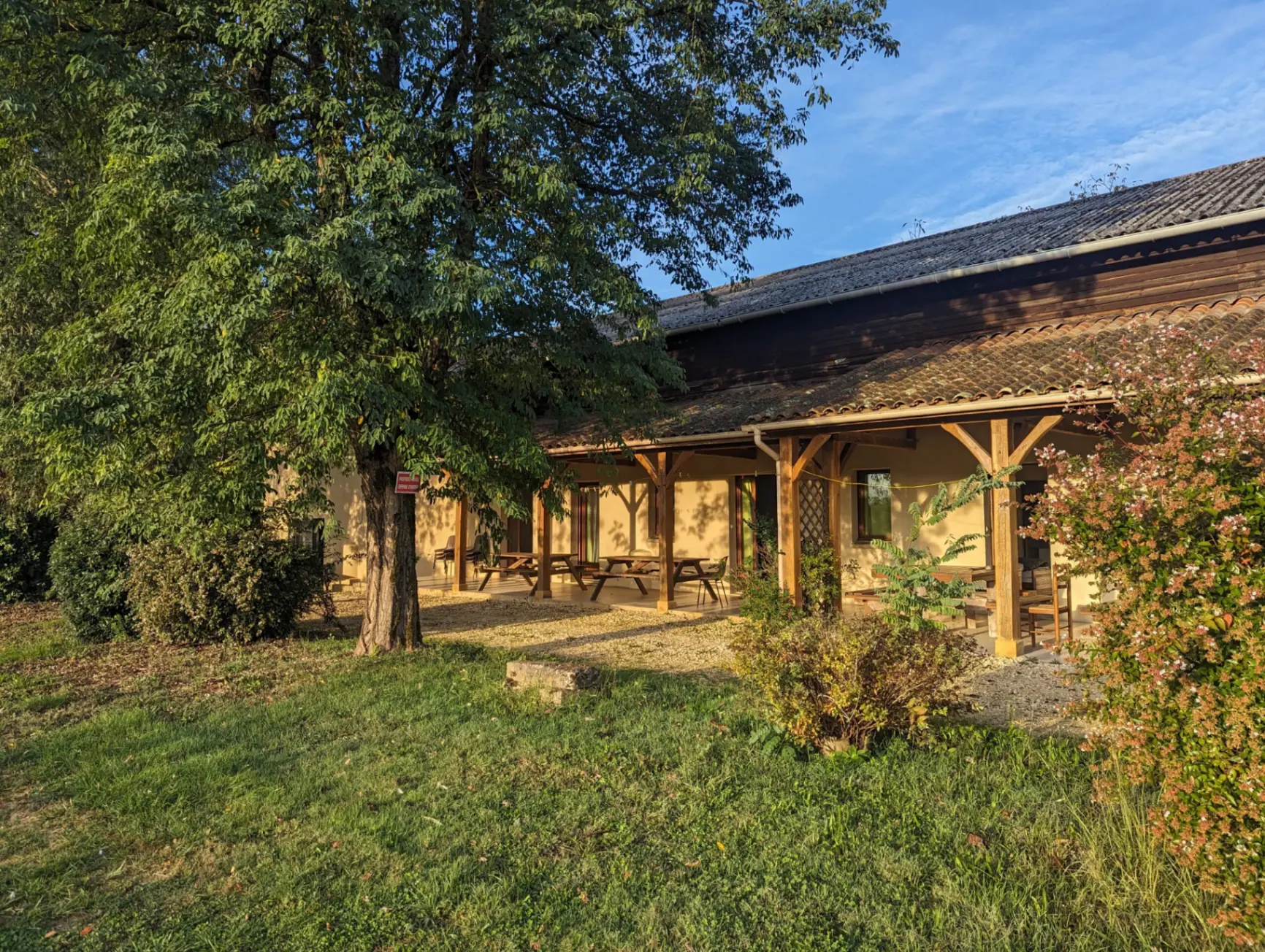 Terrasse couverte avec charpente en bois, tables de pique-nique et vegetation fleurie en Perigord