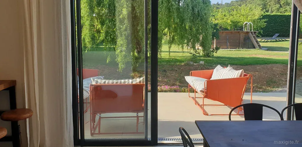 Vue depuis le séjour sur la terrasse avec canapés orange et jardin avec piscine en arrière-plan
