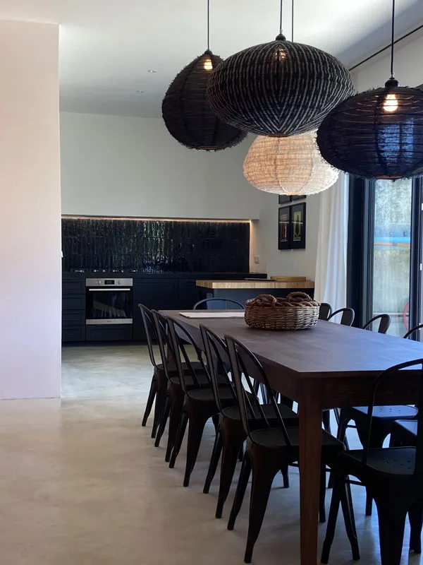 Grande salle à manger avec longue table en bois, chaises métalliques et suspensions en osier tressé