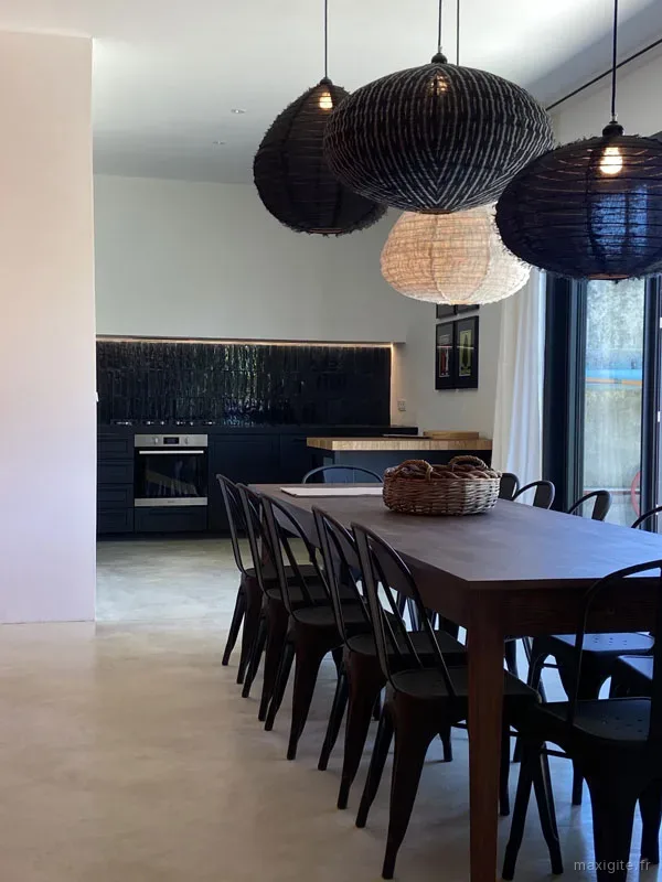 Grande salle à manger avec longue table en bois, chaises métalliques et suspensions en osier tressé