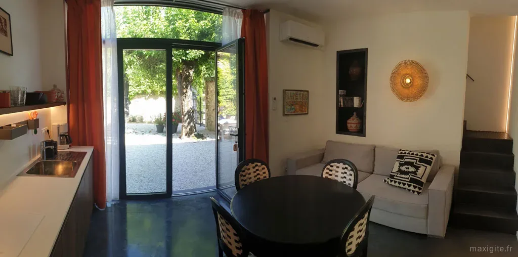 Séjour lumineux avec canapé, table ronde, kitchenette et grandes baies vitrées ouvertes sur la cour