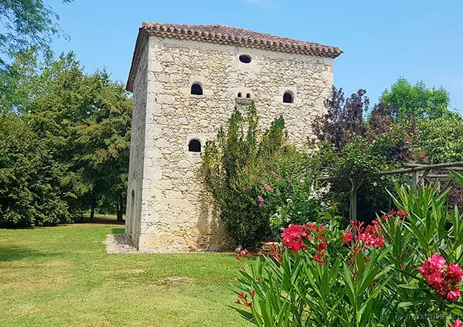 Pigeonnier en pierre avec ouvertures rondes dans le parc arboré, lauriers roses en fleur et pelouse verte au premier plan