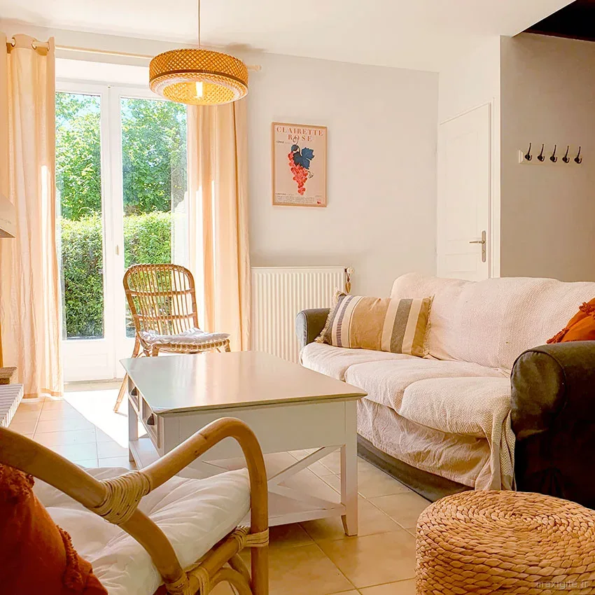 Salon lumineux avec canapé recouvert d'un jeté beige, fauteuil en rotin, table basse blanche et baie vitrée sur le jardin
