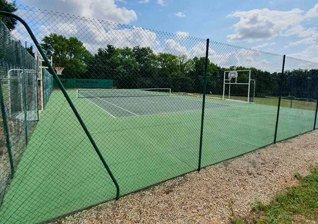 Terrain multisports clôturé avec surface synthétique verte, filet de tennis, panneaux de basket et arbres en arrière-plan